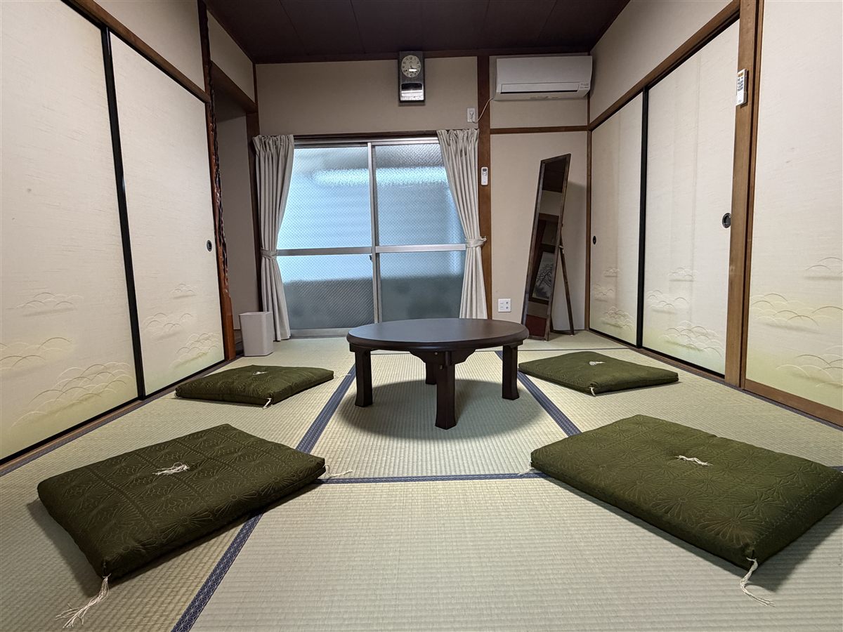 1階の和室。畳の中央に丸いちゃぶ台と4枚の座布団が置かれ、窓にはカーテンがかかっている。